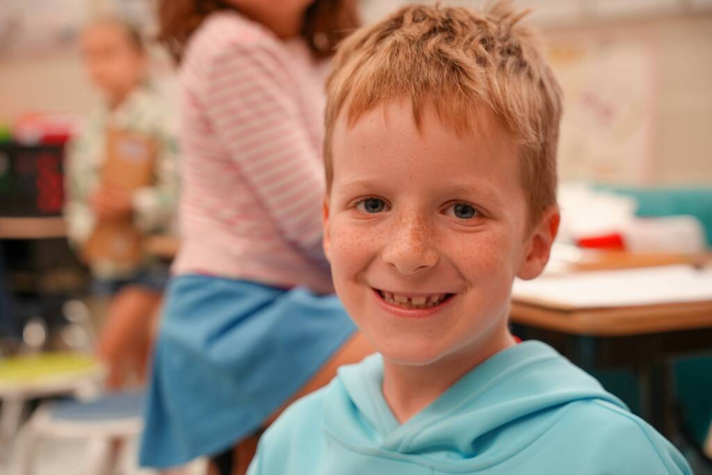 A young boy with short blonde hair smiles, wearing a blue hoodie, in a classroom with blurred background.