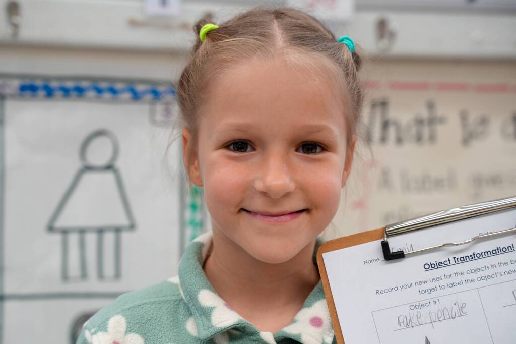 A smiling child with pigtails holds a clipboard, showing a form. A drawing of a woman is behind her.