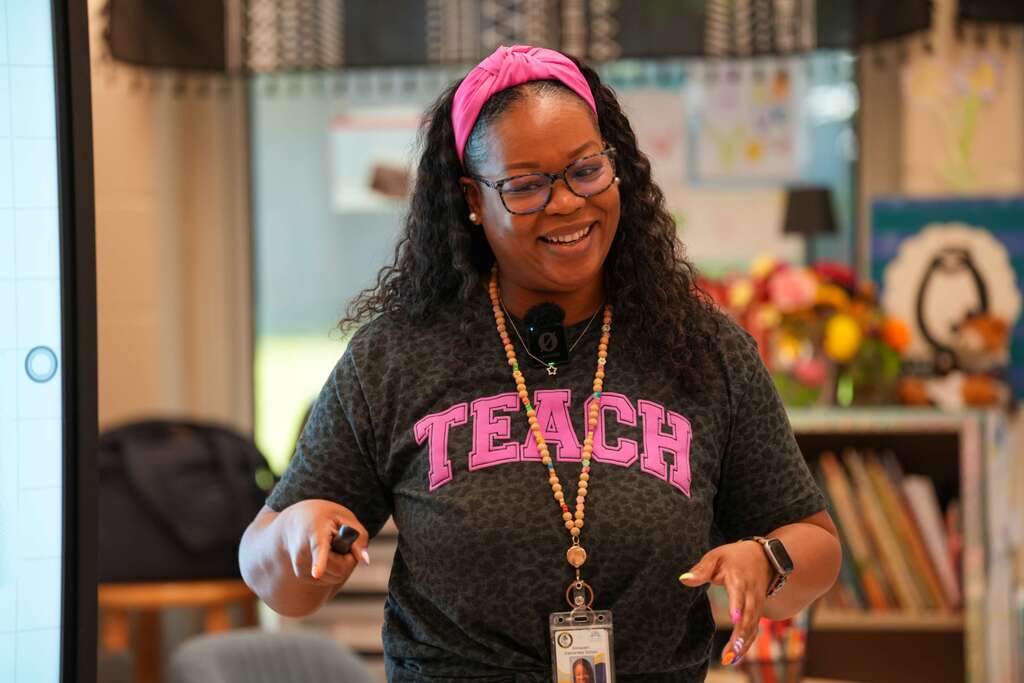 A woman in a black shirt with "TEACH" in pink letters stands in a classroom. She wears a pink headband and glasses.