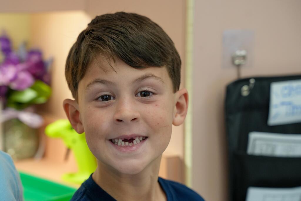 A boy with brown hair smiles and shows missing teeth. A blurry background shows a vase with flowers, a yellow chair, and a bulletin board.