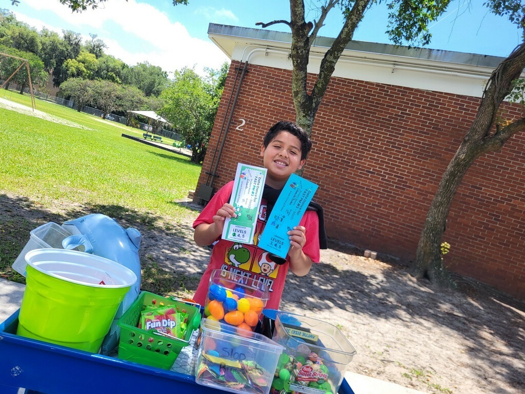 A young person stands by a cart filled with candies and a blue sign. Behind them is a brick building and green grass.