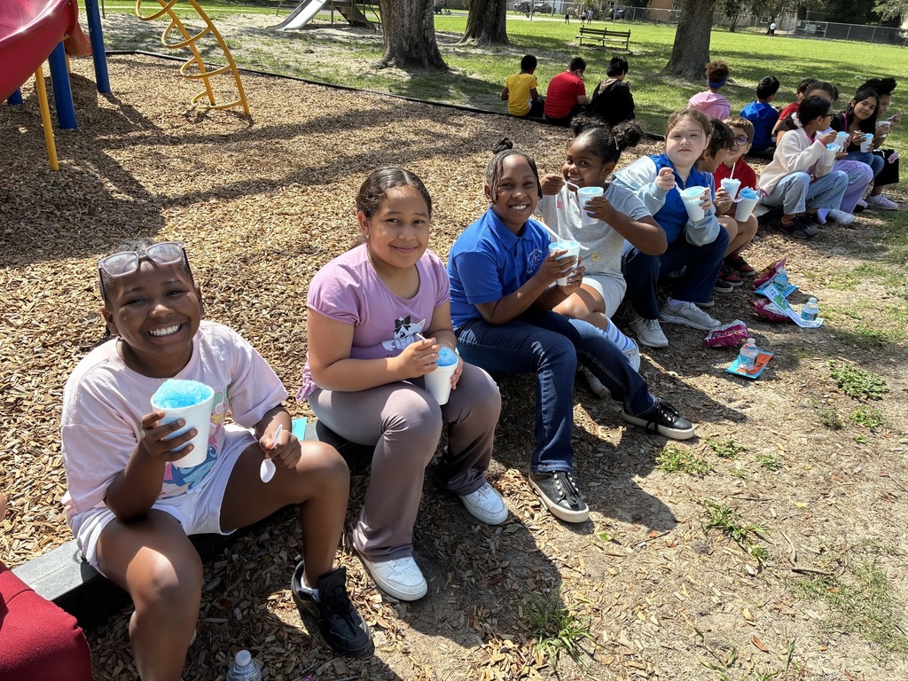 Group of children sitting in a line holding cups, some smiling. A playground is in the background.