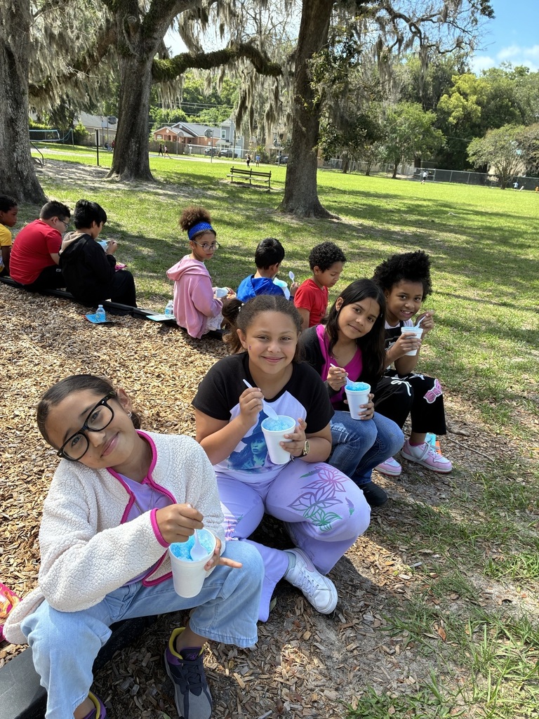 Kids sitting on a grassy area, some with cups of snow cones, under a tree.