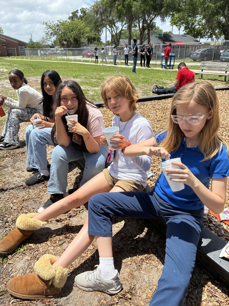Group of children sitting on ground eating snow cones. Trees, people, and buildings in background.