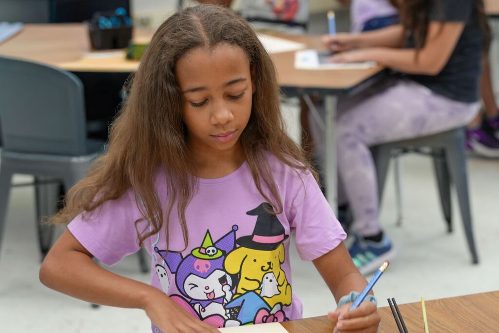 A young girl in a purple shirt with a cartoon character prints writes on a piece of paper.