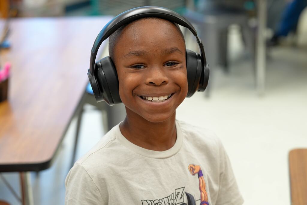 A smiling young boy with headphones on, wearing a white t-shirt, seated in a classroom with tables and chairs.