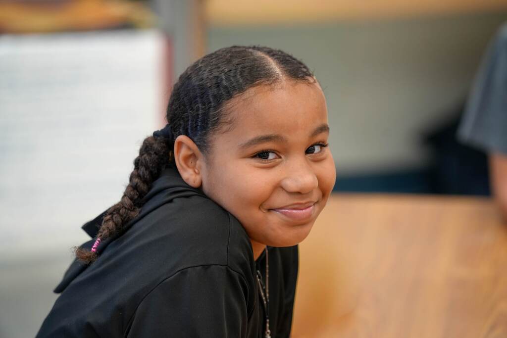 A girl with braided hair smiles, wearing a black hoodie. Background includes a wooden surface and blurred people.