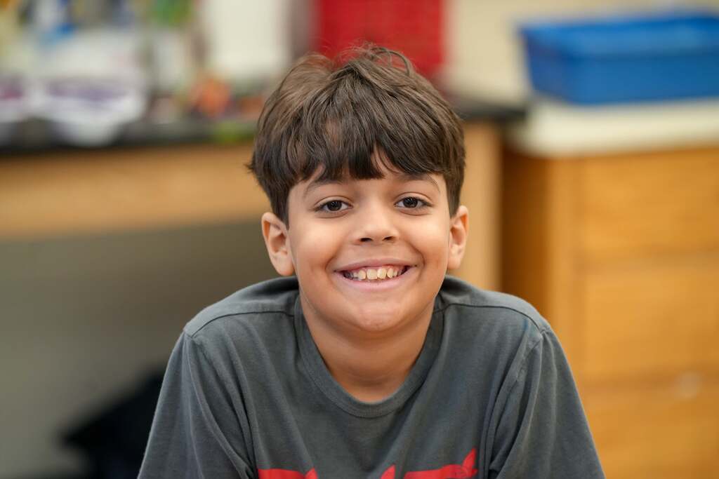 A smiling child in a gray t-shirt sits indoors. Behind the child is a wooden cabinet with a blue bin.
