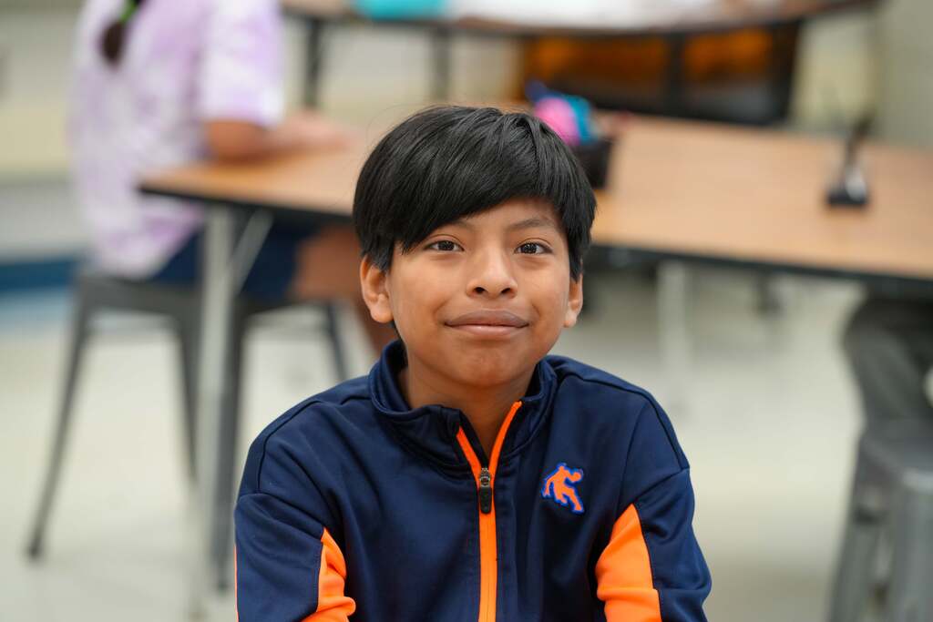 A boy in a blue and orange jacket sits in a classroom. Behind him is a desk with a blurred person.