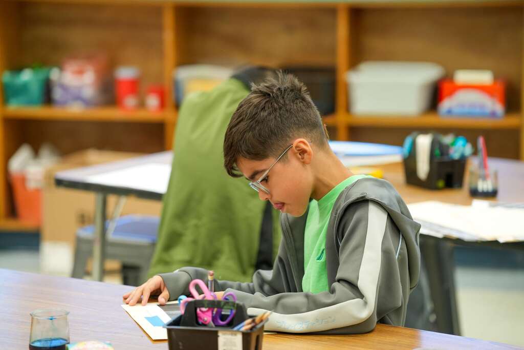A young boy wearing glasses sits at a desk in a classroom, focused on a book.