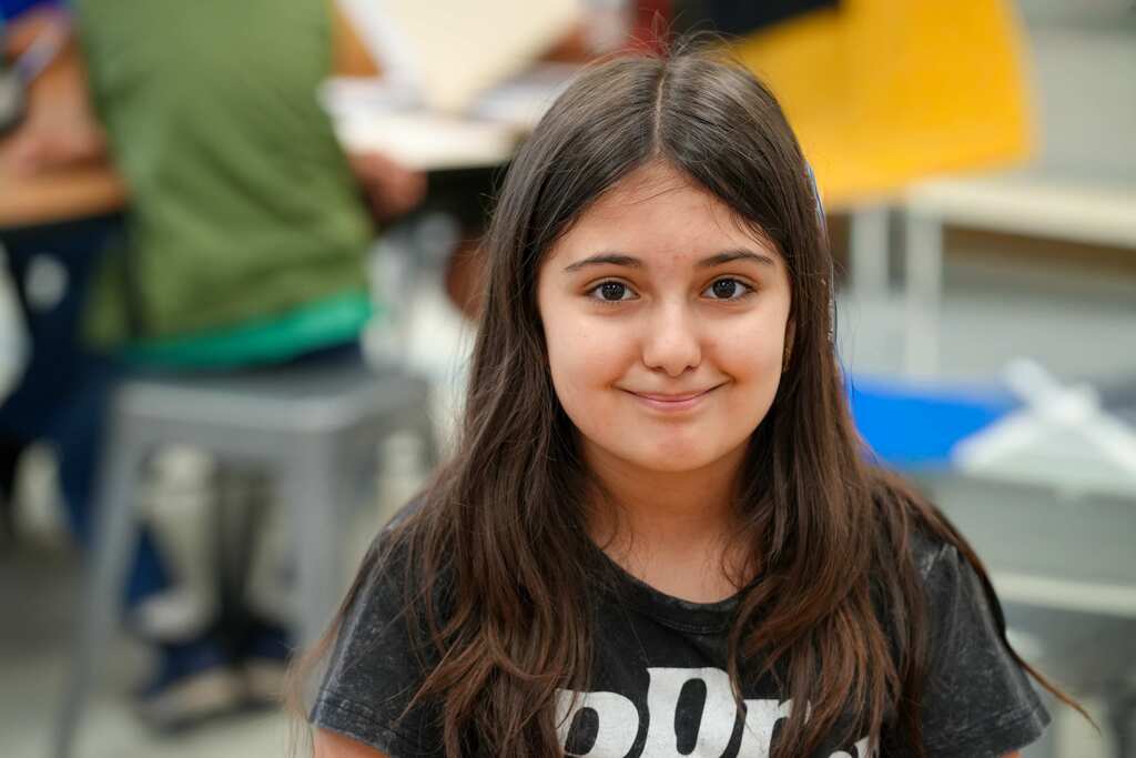 A young girl with brown hair, wearing a black t-shirt, smiles at the camera. Behind her, blurred figures work at tables.