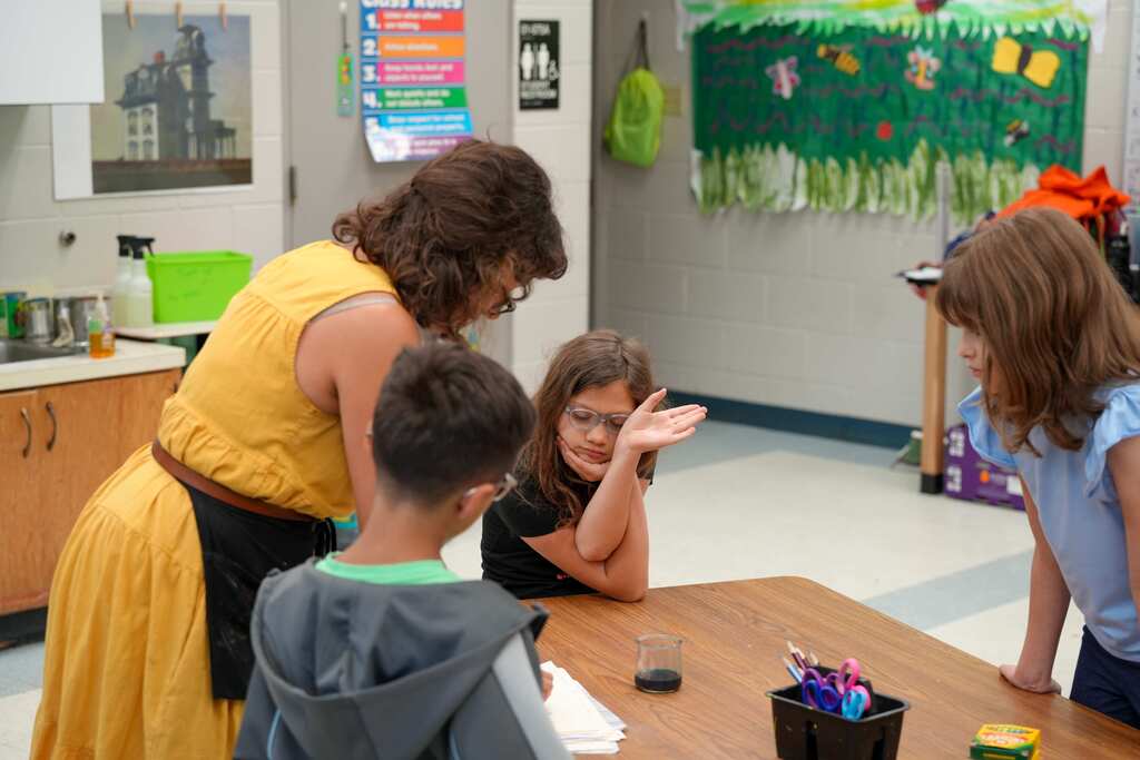 A woman in yellow stands near three kids, one seated at a table with items. Behind them, a white wall with decorations and a monitor.