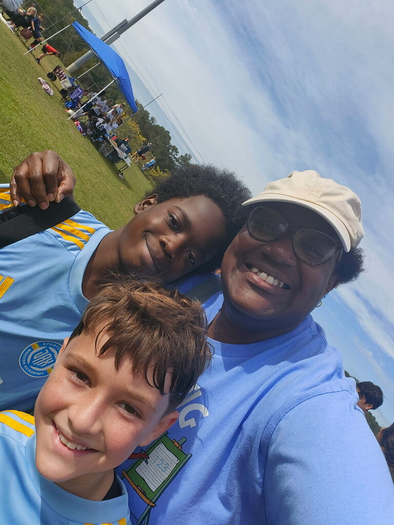 A teacher smiles at the soccer field while posing with two students in soccer gear