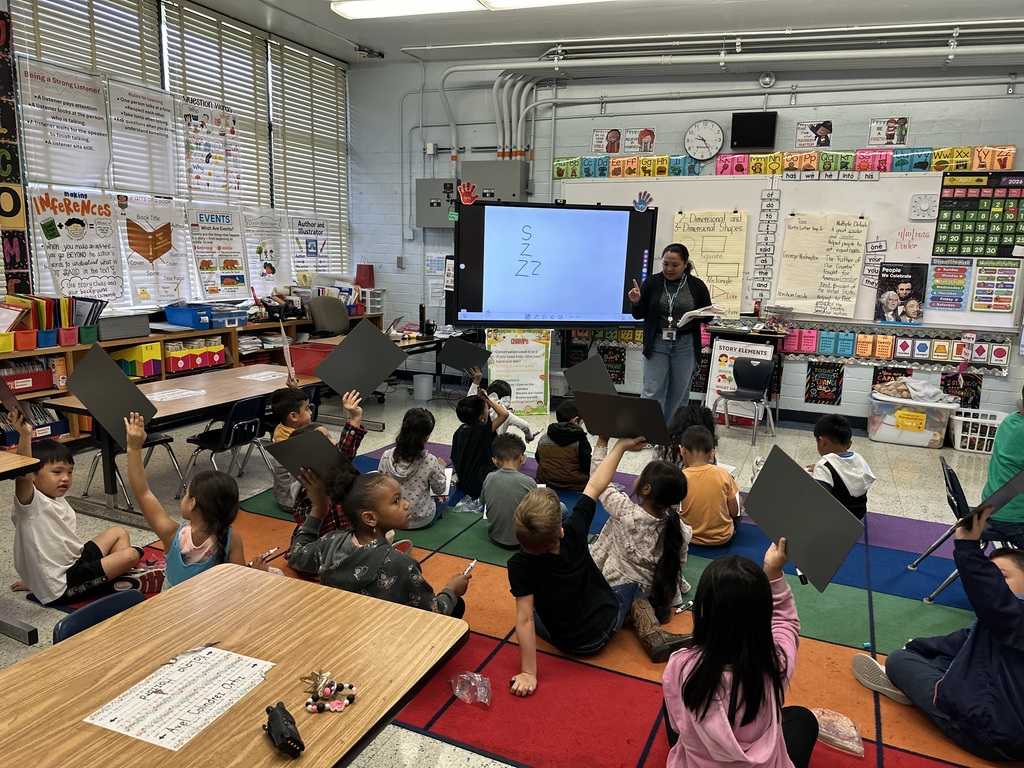 A teacher stands in a classroom instructing a group of students who are holding whiteboards
