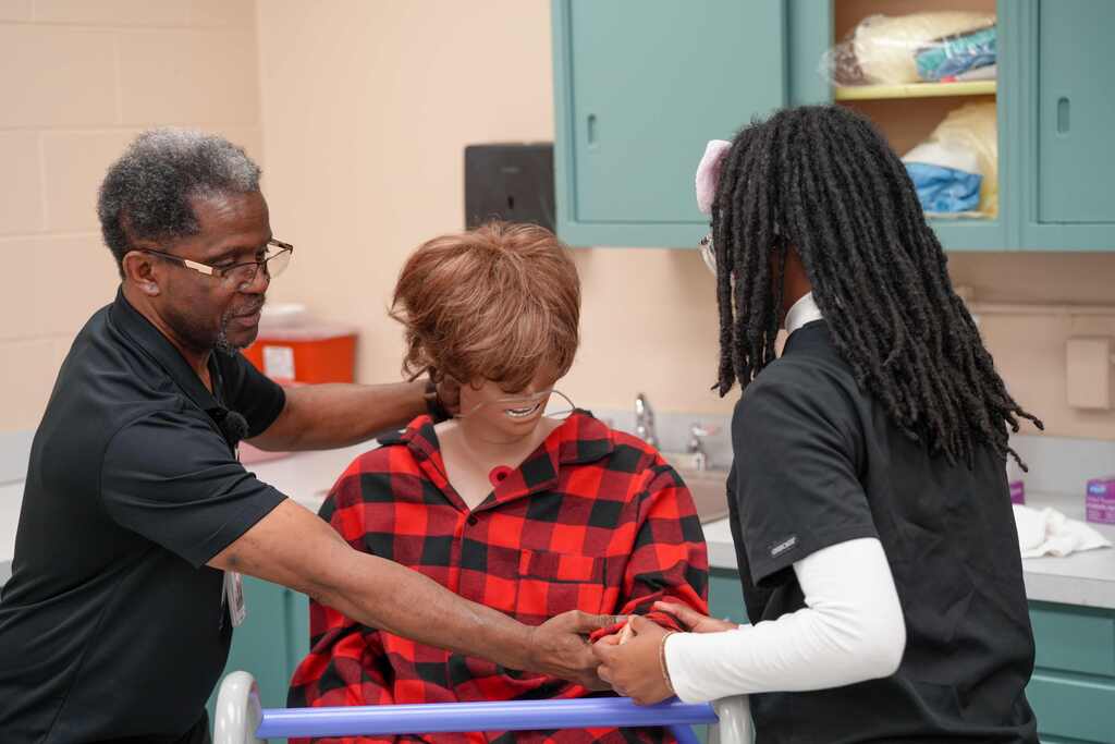 A person in a wheelchair receives assistance from two others in a kitchen with cabinets.