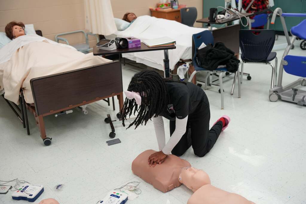 A person practices CPR on a mannequin in a medical training room with beds and chairs around.