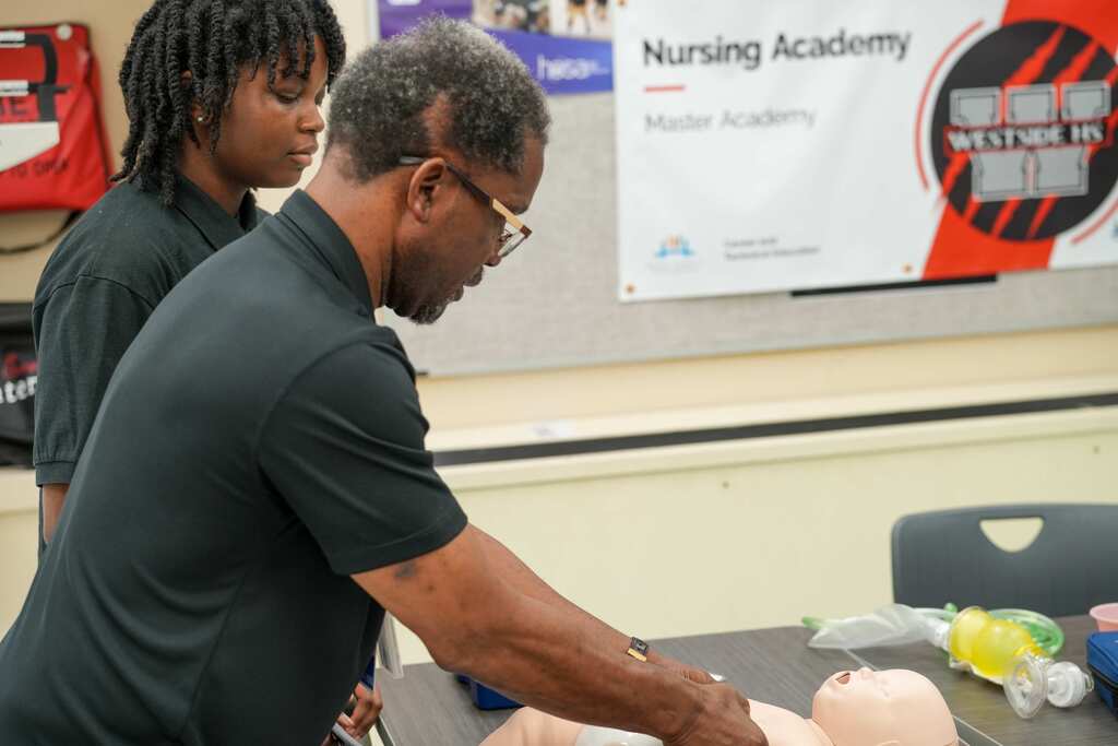 Man with glasses and a woman with braids in a room with a Nursing Academy banner and a dummy.