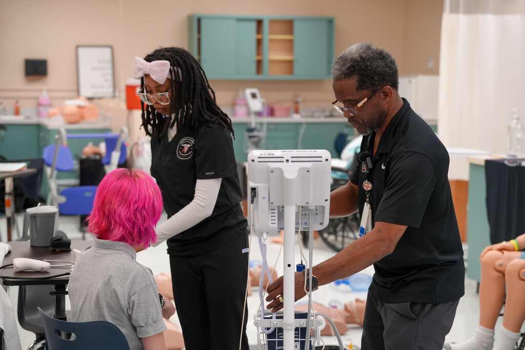 Two people in black scrubs work with a mannequin in a medical training room with a pink-haired student.