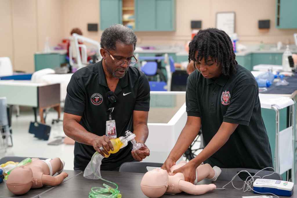 Two people in black shirts work on a CPR training dummy at a table. A room with desks and other equipment is behind them.