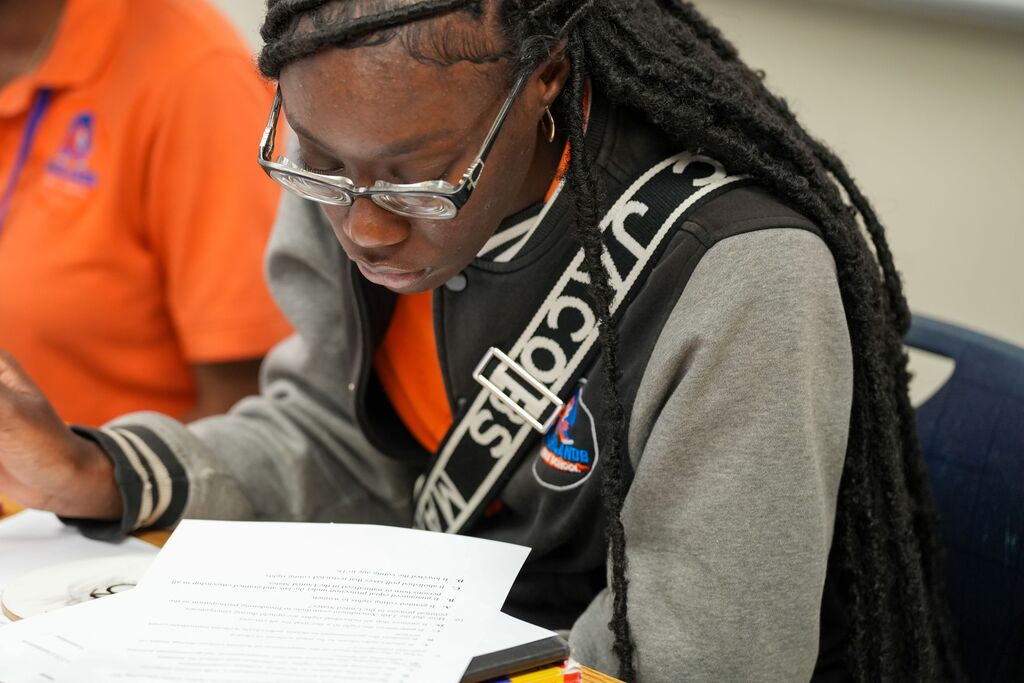 A student sits with papers, wearing glasses and a gray and orange jacket with a NASA logo.