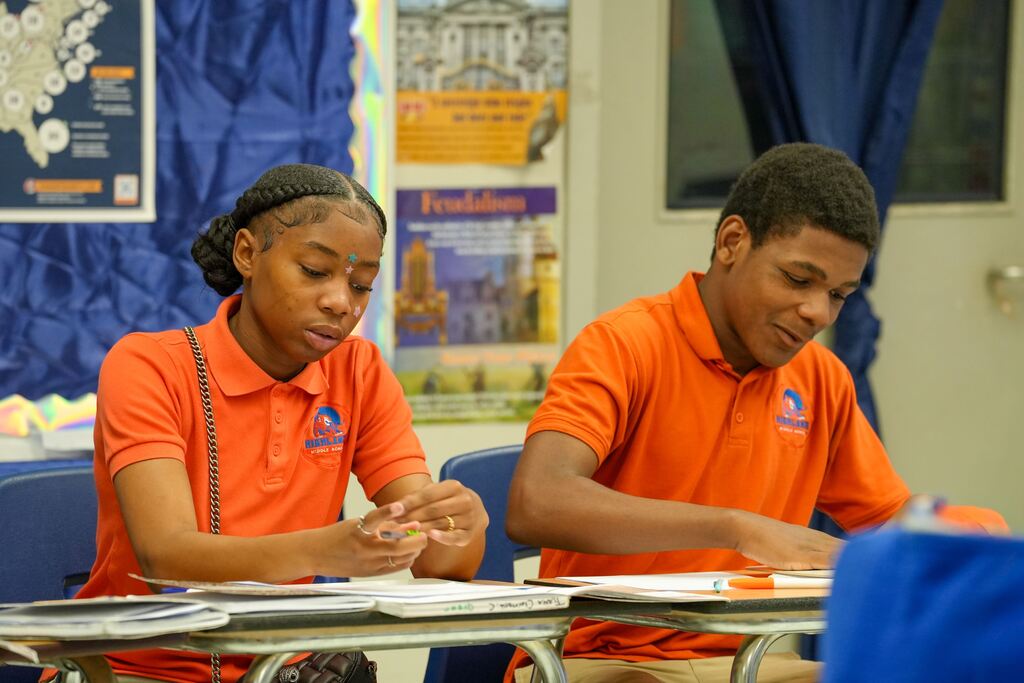 Two students in orange shirts sit at a desk. They focus on papers and a blue curtain behind them.