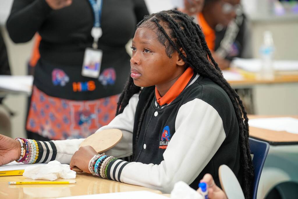 A student with long braids and a white-black jacket sits at a desk, holding a pencil.
