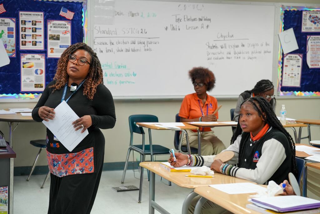 Woman standing with paper in hand in front of class. Students seated at desks, one writing.