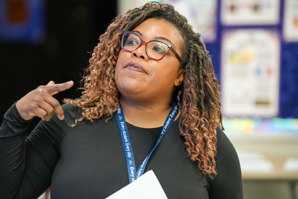 A woman with curly hair wearing glasses and a black shirt stands in a classroom, gesturing with her hand.