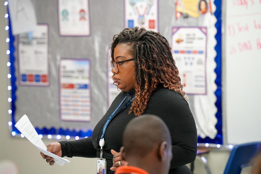 Woman with curly hair speaks to a man in a classroom with a bulletin board behind her.