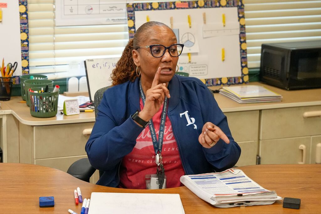 Person seated at desk in classroom, pointing finger, wearing glasses and blue jacket, surrounded by papers, pens, and other items.