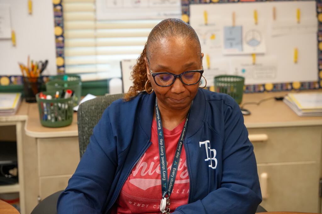 A woman with short hair wearing glasses and a blue jacket sits at a desk with a red shirt.