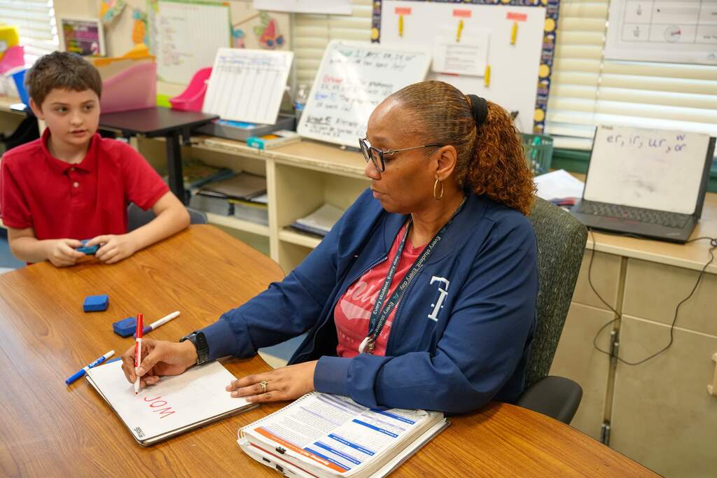 A woman in a blue jacket works at a desk while a child in a red shirt sits nearby.