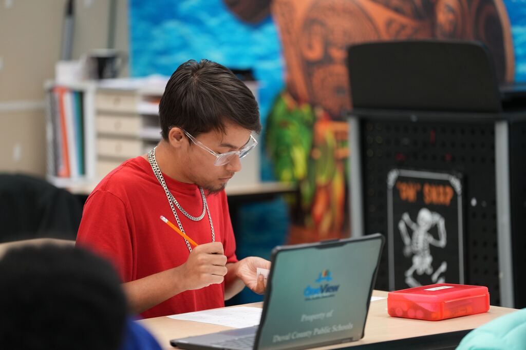 A man wearing a red T-shirt and glasses sits at a desk with a laptop, writing.