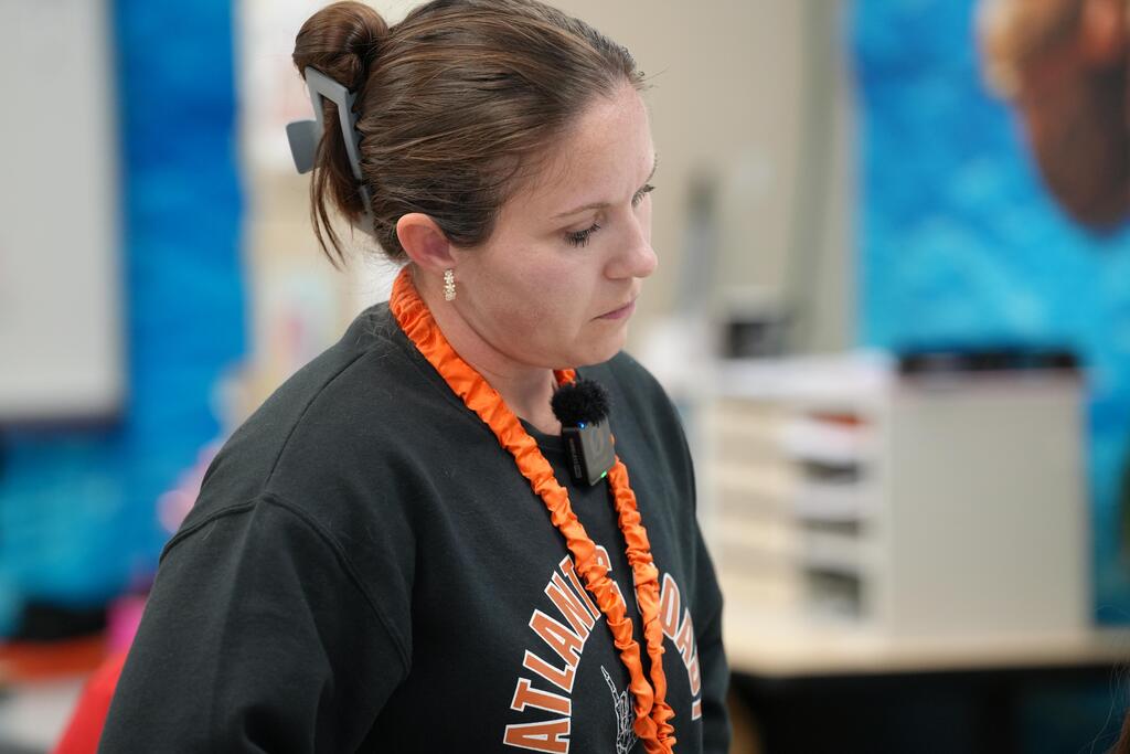 Woman wearing a black shirt and orange necklace standing in a classroom, looking down.