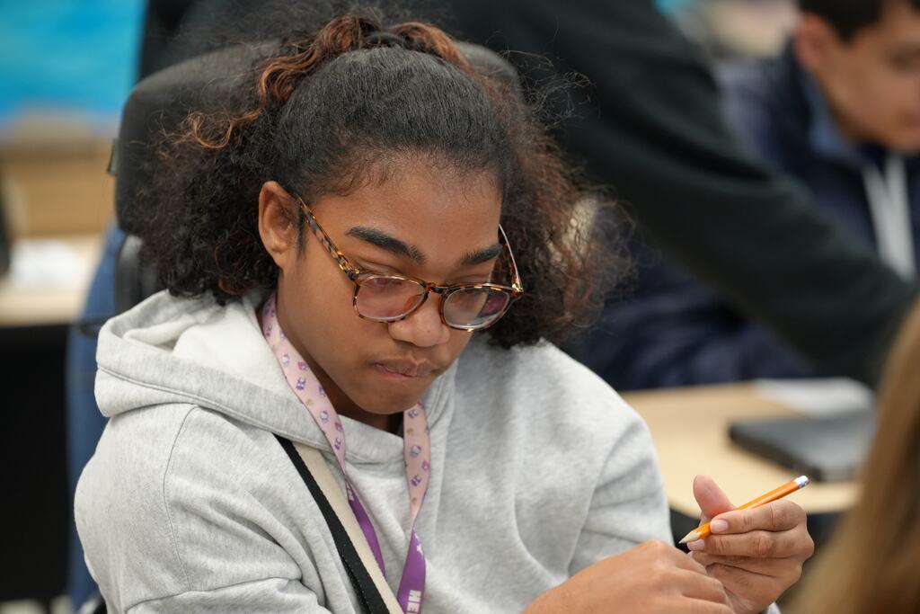 A student wearing glasses focuses on writing in a classroom, seated in a chair. A backpack rests on the floor.