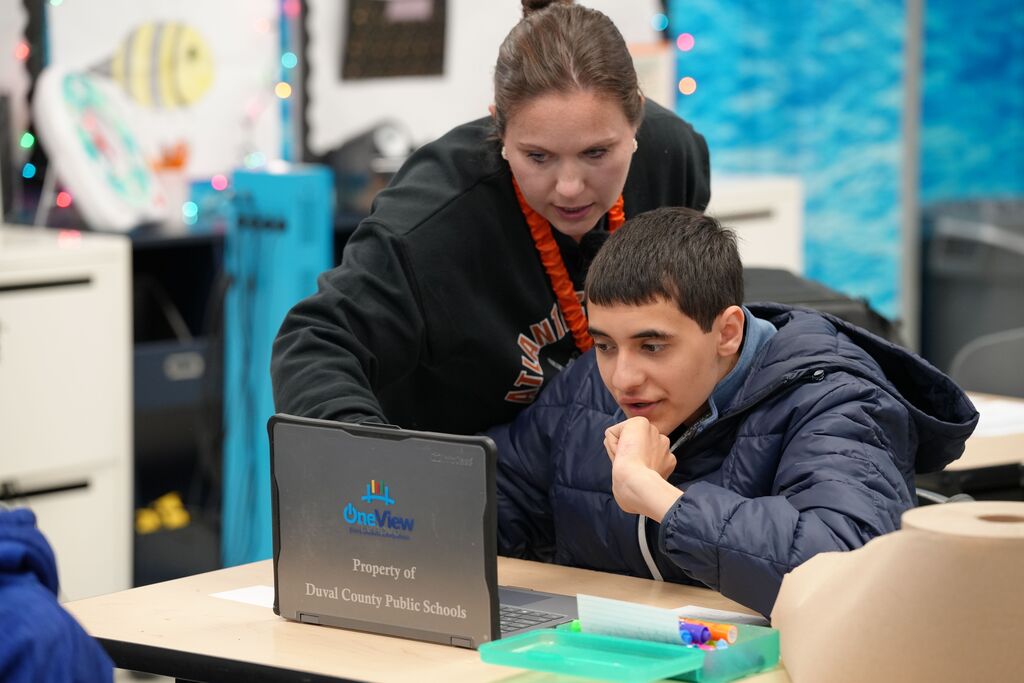 Two students work on a laptop. A woman leans over a boy wearing a blue jacket. Bright lights reflect on the wall behind them.