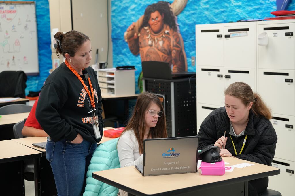 Three women in a classroom; one seated at a desk with a laptop, another standing nearby, and a third in the background.