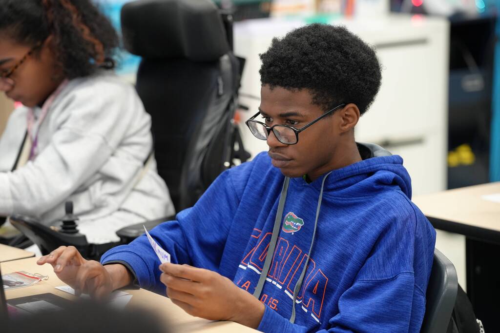 Two students in a classroom, one in a blue hoodie with glasses, focusing on a task.