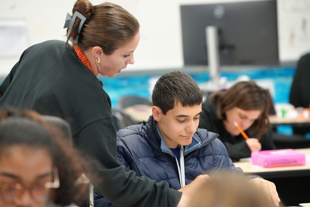 A teacher assists a student in a classroom. Behind them, another student writes at a desk.