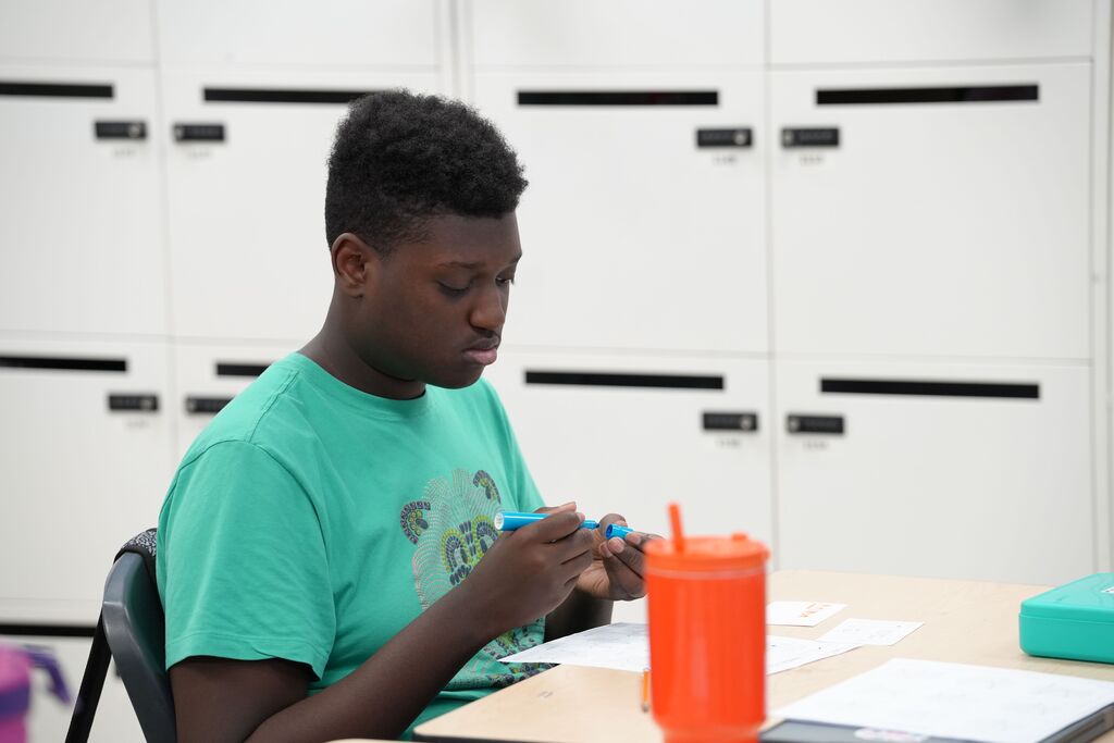 A person in a green shirt works on a craft project, seated at a desk with white cabinets in the background.