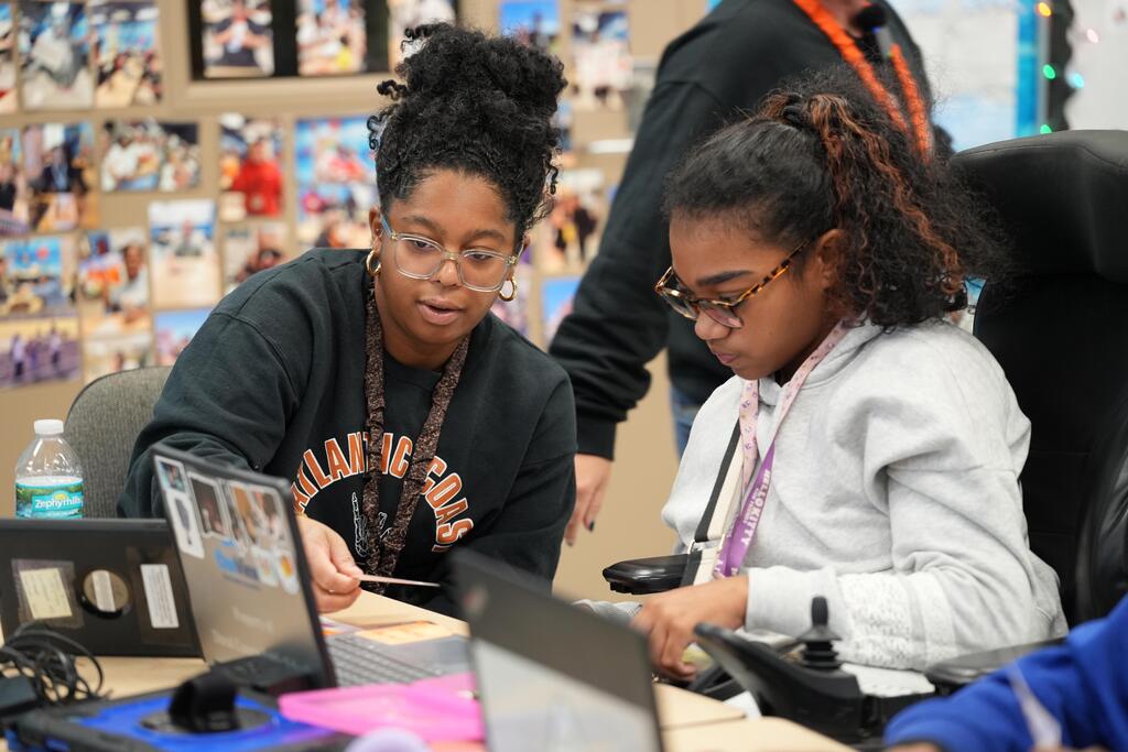 Two young women in glasses sit at a table. One uses a computer while the other works on a paper.