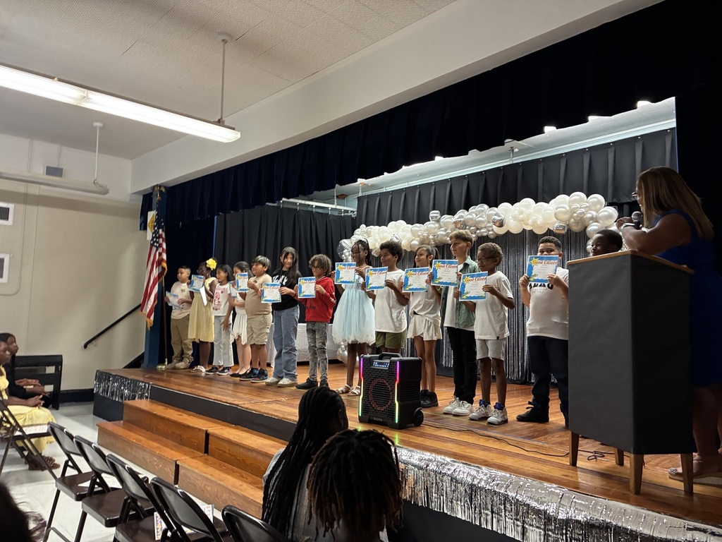 A group of children holding certificates stand on a stage with a podium and a flag.