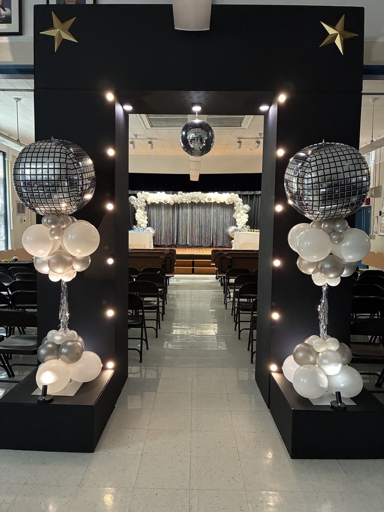 Black pillars with disco balls and balloons leading to an aisle with chairs and a stage.