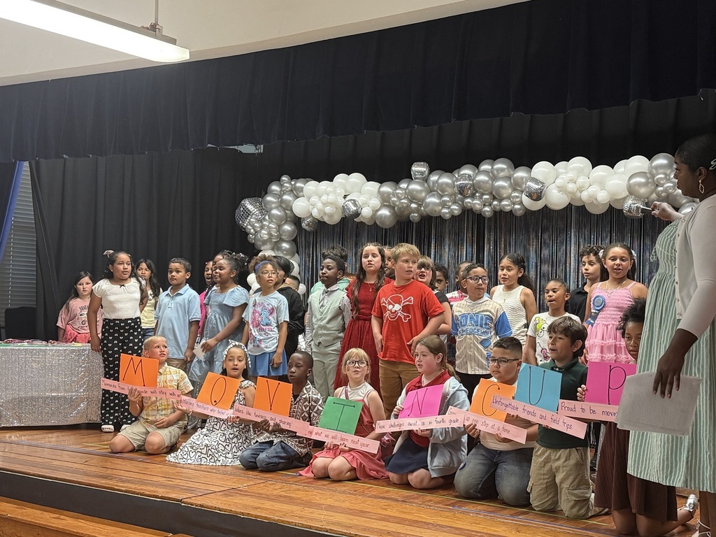 A group of children wearing costumes and holding signs on a stage with balloons and a backdrop.