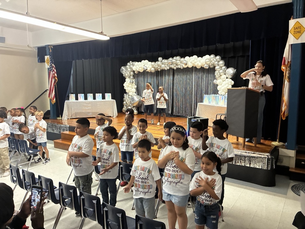 A group of children in white shirts on a stage, performing in front of an audience.