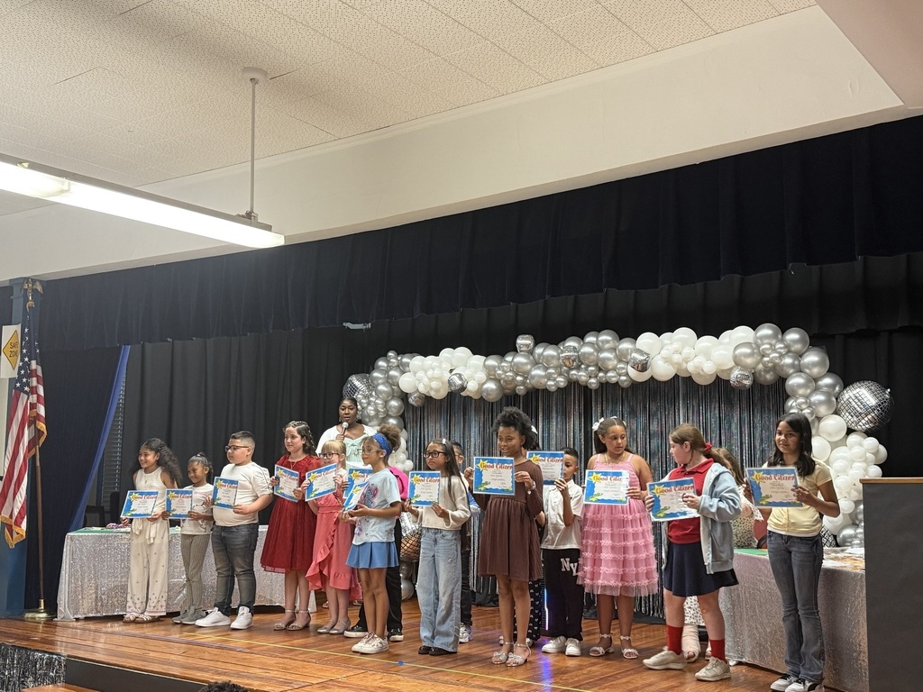 Several children on a stage holding certificates, standing in front of a backdrop with balloons