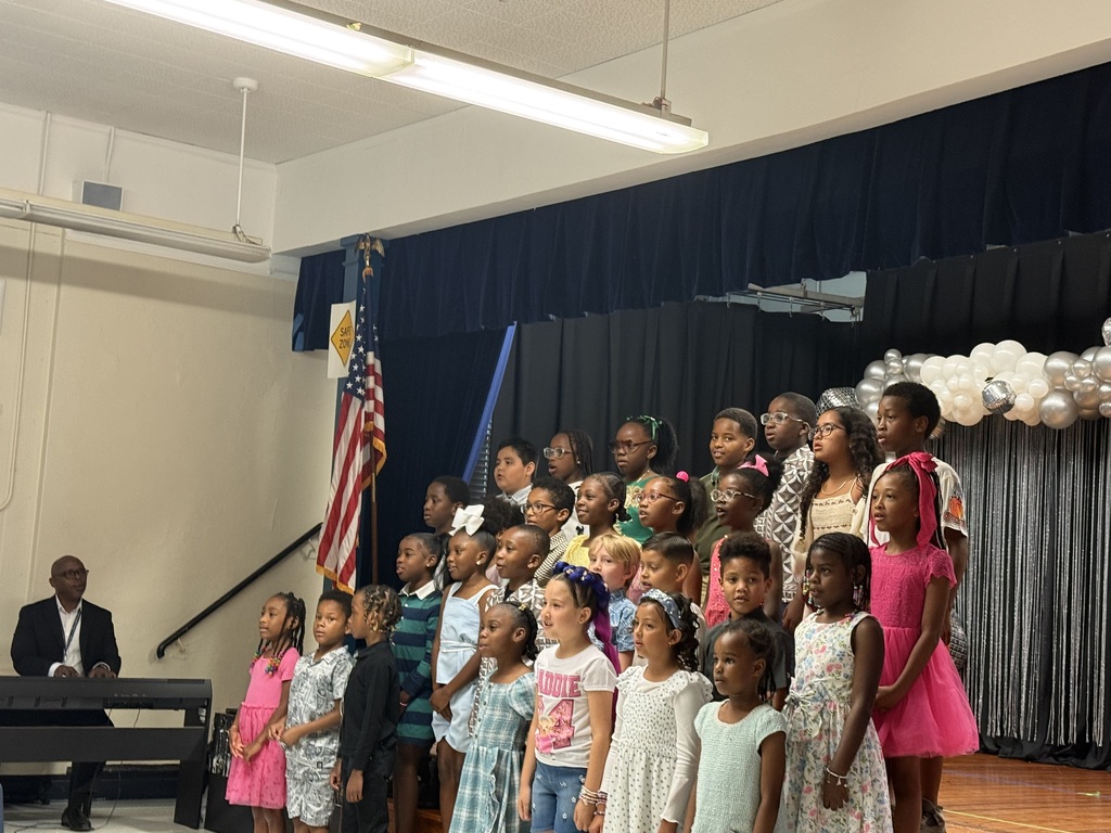A group of children dressed in various outfits stand on a stage with a flag. An adult is seated at a piano.