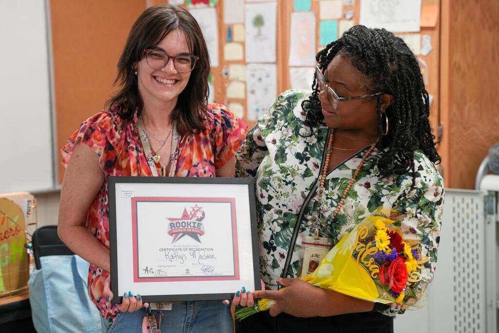 A woman holding a framed award with flowers, assisted by another woman in a classroom.
