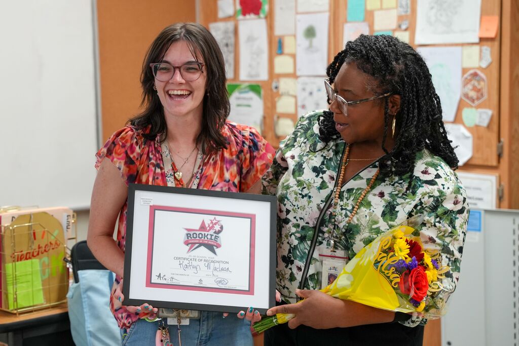 Two women in a classroom holding a framed award and flowers. The award has text and images.