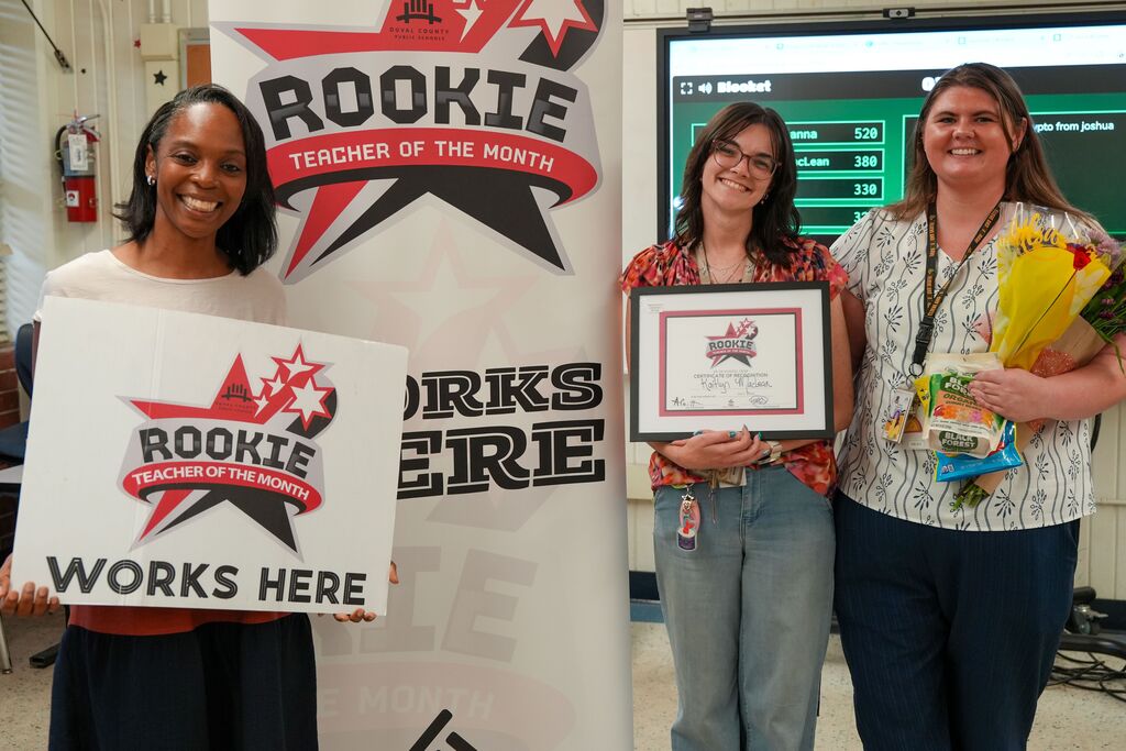 Three women stand behind a banner, two holding signs and one holding a framed certificate.